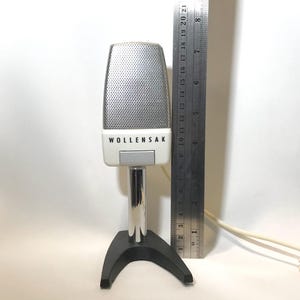 May include: A Wollensak microphone with a silver mesh grille and white casing, mounted on a chrome stand with a black base. A metal ruler is positioned next to the microphone, showing a height of approximately 9 inches from the base to the top.