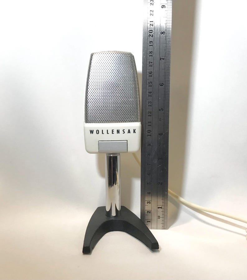 May include: A Wollensak microphone with a silver mesh grille and white casing, mounted on a chrome stand with a black base. A metal ruler is positioned next to the microphone, showing a height of approximately 9 inches from the base to the top.