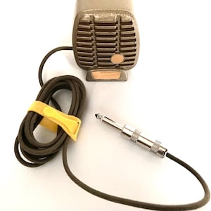 Puede incluir: Un micr&oacute;fono de estilo vintage color bronce con una rejilla rectangular y un peque&ntilde;o acento circular. Cuenta con un cable largo de color marr&oacute;n oscuro enrollado y asegurado con una tira de velcro amarilla brillante, que termina en un conector jack de metal plateado.