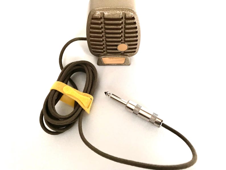 Puede incluir: Un micr&oacute;fono de estilo vintage color bronce con una rejilla rectangular y un peque&ntilde;o acento circular. Cuenta con un cable largo de color marr&oacute;n oscuro enrollado y asegurado con una tira de velcro amarilla brillante, que termina en un conector jack de metal plateado.