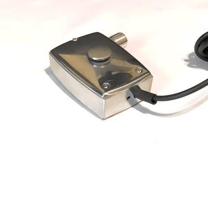 Puede incluir: Una caja de control rectangular de metal plateado pulido con un &uacute;nico bot&oacute;n circular en la superficie superior, asegurada por cuatro tornillos en las esquinas. Un cable el&eacute;ctrico negro sale del lateral y se observa un peque&ntilde;o puerto cil&iacute;ndrico.