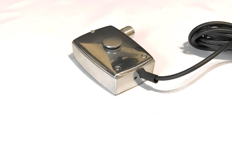 Puede incluir: Una caja de control rectangular de metal plateado pulido con un &uacute;nico bot&oacute;n circular en la superficie superior, asegurada por cuatro tornillos en las esquinas. Un cable el&eacute;ctrico negro sale del lateral y se observa un peque&ntilde;o puerto cil&iacute;ndrico.