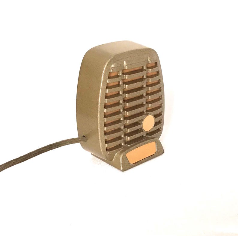 Puede incluir: Un micr&oacute;fono de estilo vintage en tonos dorados con un dise&ntilde;o de rejilla rectangular, lamas horizontales, un acento circular y una peque&ntilde;a placa base rectangular. Un cable fino recubierto de tela oscura sale del lateral del dispositivo.