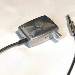 Puede incluir: Un dispositivo rectangular de metal plateado con un bot&oacute;n circular en la parte superior y una perilla de control giratoria lateral. Un cable negro sale de la unidad y termina en un conector jack de audio est&aacute;ndar de 6,35 mm.