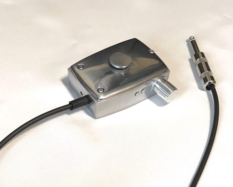 Puede incluir: Un dispositivo rectangular de metal plateado con un bot&oacute;n circular en la parte superior y una perilla de control giratoria lateral. Un cable negro sale de la unidad y termina en un conector jack de audio est&aacute;ndar de 6,35 mm.