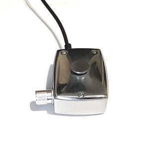 Puede incluir: Una caja de control rectangular de metal cromado pulido que presenta un bot&oacute;n pulsador circular en la superficie superior, una perilla de ajuste cil&iacute;ndrica estriada en el lateral y un cable el&eacute;ctrico negro que sale del borde superior.