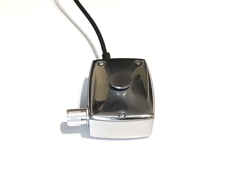 Puede incluir: Una caja de control rectangular de metal cromado pulido que presenta un bot&oacute;n pulsador circular en la superficie superior, una perilla de ajuste cil&iacute;ndrica estriada en el lateral y un cable el&eacute;ctrico negro que sale del borde superior.