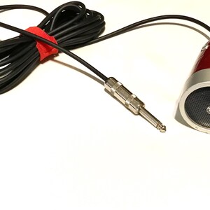 May include: A red and silver microphone with a black mesh grille and a long black cable. The cable features a silver-colored 1/4-inch jack plug and is secured with a small red hook-and-loop fastener strap to keep the cord neatly coiled.