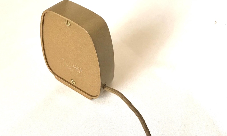 Puede incluir: Un dispositivo el&eacute;ctrico de color marr&oacute;n claro, de forma cuadrada con esquinas redondeadas y un acabado mate liso. El panel trasero est&aacute; asegurado con dos tornillos visibles y un cable el&eacute;ctrico fino de color marr&oacute;n claro sale del lado inferior derecho.
