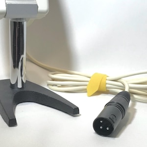 May include: A silver and grey microphone mounted on a chrome-plated cylindrical post with a black V-shaped base. The microphone includes a long, off-white cable secured with a yellow tie and terminates in a black three-pin XLR connector plug.