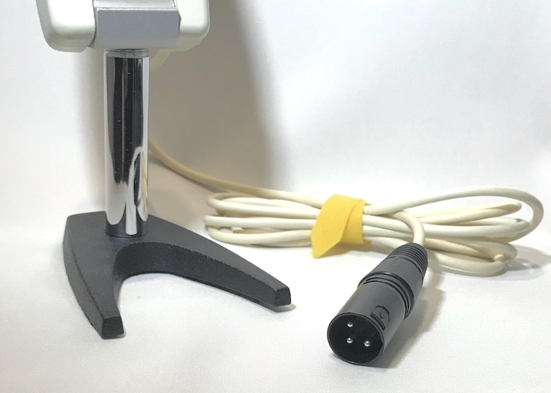 May include: A silver and grey microphone mounted on a chrome-plated cylindrical post with a black V-shaped base. The microphone includes a long, off-white cable secured with a yellow tie and terminates in a black three-pin XLR connector plug.