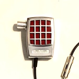 Puede incluir: Un micr&oacute;fono plateado con una cabeza rectangular que presenta un patr&oacute;n de rejilla rojo. La base del micr&oacute;fono tiene la etiqueta "MODEL 715 CERAMIC". Un cable negro se extiende desde la parte inferior, conect&aacute;ndose a un conector jack de audio.