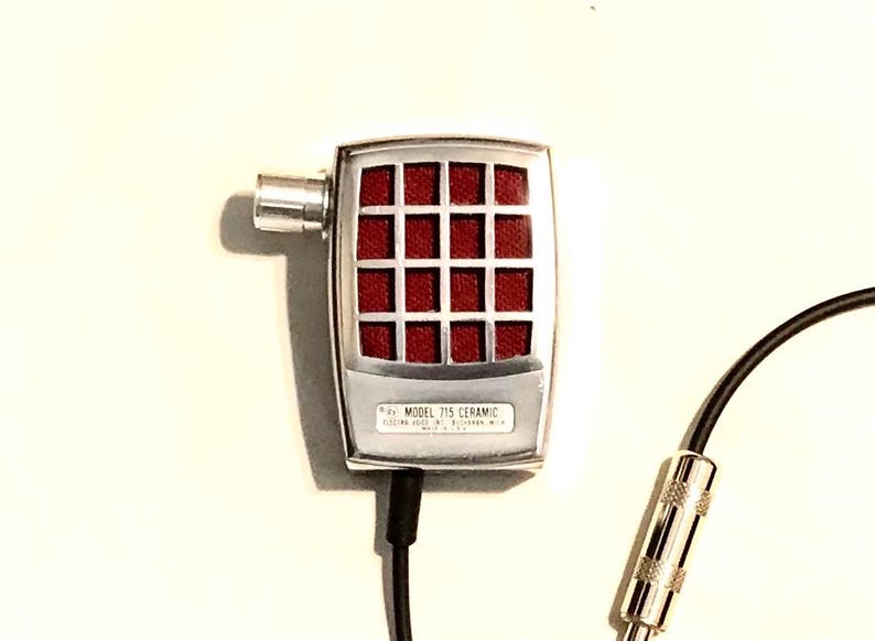 Puede incluir: Un micr&oacute;fono plateado con una cabeza rectangular que presenta un patr&oacute;n de rejilla rojo. La base del micr&oacute;fono tiene la etiqueta "MODEL 715 CERAMIC". Un cable negro se extiende desde la parte inferior, conect&aacute;ndose a un conector jack de audio.