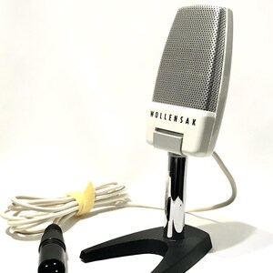 May include: A vintage Wollensak microphone featuring a white casing with a silver perforated grille. The microphone is mounted on a chrome-plated vertical stand attached to a black V-shaped base. A white cable with a black three-pin connector is attached.