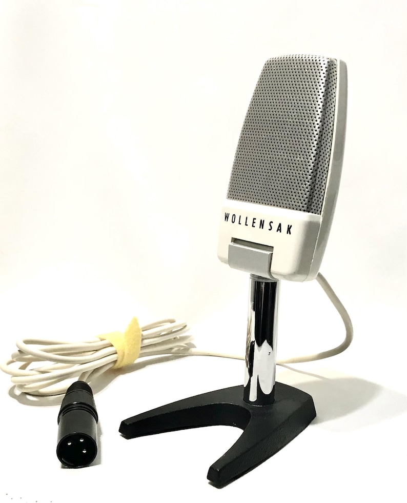 May include: A vintage Wollensak microphone featuring a white casing with a silver perforated grille. The microphone is mounted on a chrome-plated vertical stand attached to a black V-shaped base. A white cable with a black three-pin connector is attached.