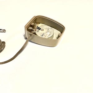 Puede incluir: Una carcasa de micr&oacute;fono de contacto rectangular de color beige con la parte posterior abierta que revela un elemento transductor circular plateado. Un cable beige fino recubierto de tela sale de la carcasa y termina en un jack de audio de 6,35 mm.