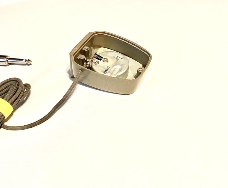 Puede incluir: Una carcasa de micr&oacute;fono de contacto rectangular de color beige con la parte posterior abierta que revela un elemento transductor circular plateado. Un cable beige fino recubierto de tela sale de la carcasa y termina en un jack de audio de 6,35 mm.