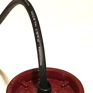 May include: A black audio cable with white text reading MOGAMI PART No.2333 connected to a circular, dark red plastic component. The cable curves upward from the center of the red base, which features a molded, ridged interior structure.
