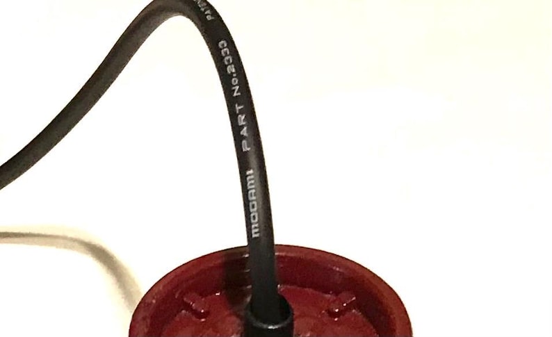 May include: A black audio cable with white text reading MOGAMI PART No.2333 connected to a circular, dark red plastic component. The cable curves upward from the center of the red base, which features a molded, ridged interior structure.