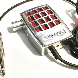Puede incluir: Un micr&oacute;fono Electro-Voice Model 715 Ceramic plateado con rejilla de malla roja, perilla de volumen lateral y cable negro con conector jack cromado de 6,35 mm. La carcasa met&aacute;lica presenta el texto Model 715 Ceramic Electro-Voice Inc.