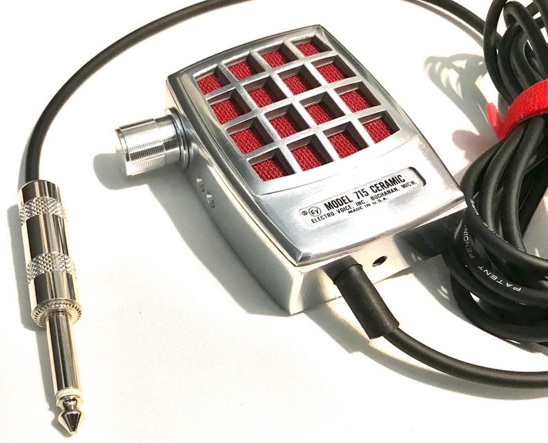 Puede incluir: Un micr&oacute;fono Electro-Voice Model 715 Ceramic plateado con rejilla de malla roja, perilla de volumen lateral y cable negro con conector jack cromado de 6,35 mm. La carcasa met&aacute;lica presenta el texto Model 715 Ceramic Electro-Voice Inc.