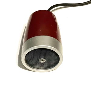 May include: A vintage-style microphone with a deep red, tapered body and a silver-toned metallic base. The front features a circular black mesh grille with a small, centered circular logo. A black electrical cord extends from the top of the unit.