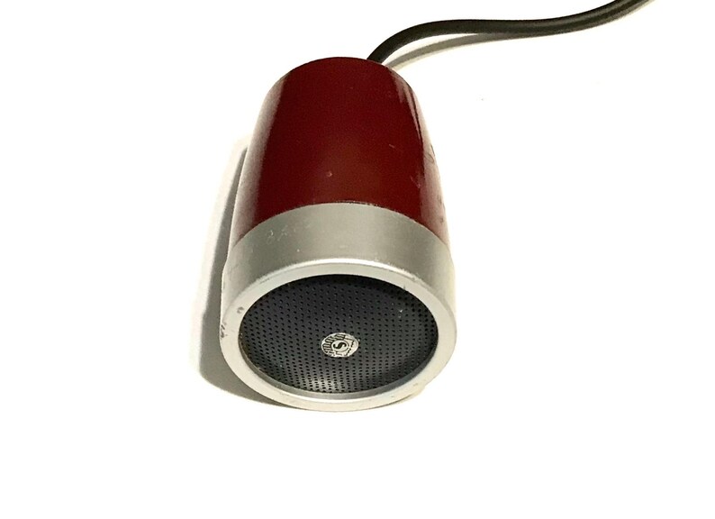 May include: A vintage-style microphone with a deep red, tapered body and a silver-toned metallic base. The front features a circular black mesh grille with a small, centered circular logo. A black electrical cord extends from the top of the unit.
