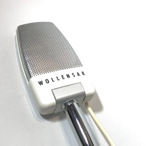 May include: A vintage Wollensak microphone featuring a rectangular silver metal grille with a fine perforated texture. The base is white plastic with the brand name Wollensak printed in black letters, connected to a polished chrome metal stand.