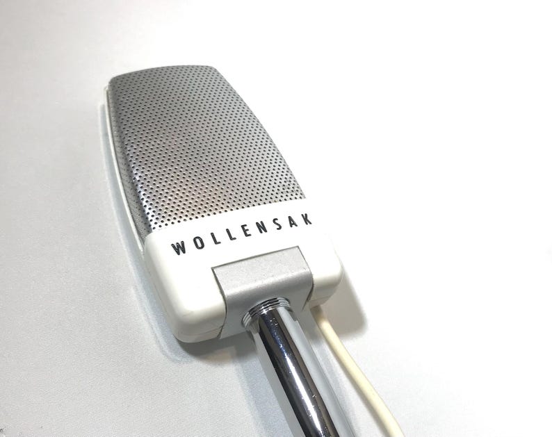 May include: A vintage Wollensak microphone featuring a rectangular silver metal grille with a fine perforated texture. The base is white plastic with the brand name Wollensak printed in black letters, connected to a polished chrome metal stand.