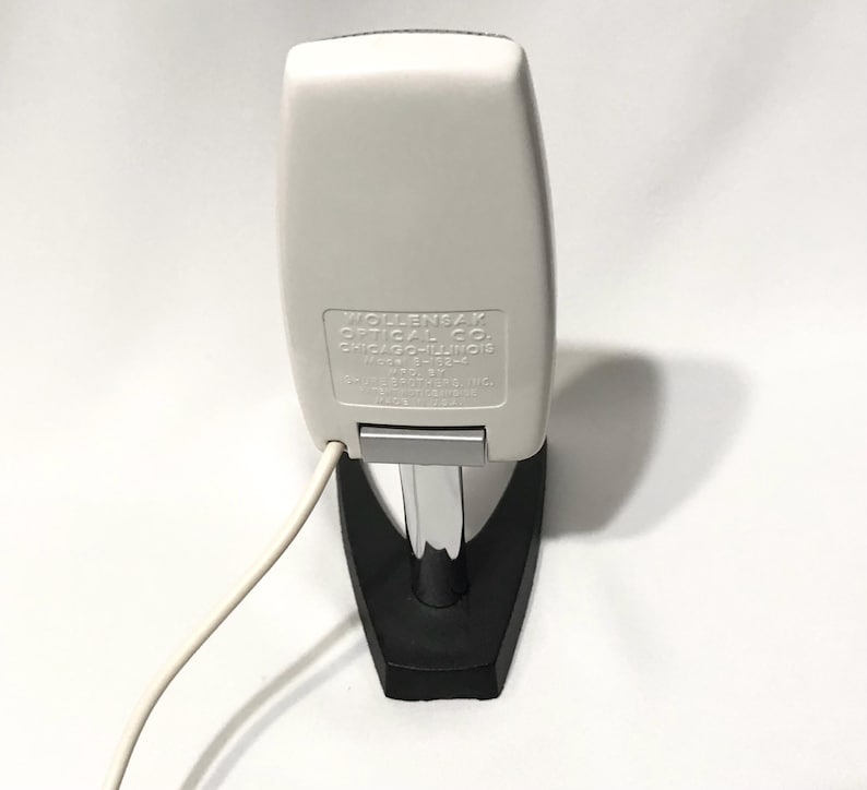 May include: A white Wollensak Optical Co. device with a chrome stand and black base. The white casing features embossed text: WOLLENSAK OPTICAL CO. CHICAGO-ILLINOIS MODEL 8H32-A MFD BY REVERE CAMERA CO. MADE IN U.S.A. A white power cord extends from the side.