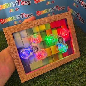 May include: Two light-up fidget spinners with red, green, and blue lights are displayed inside a wooden frame. The background features a colorful mosaic pattern. The frame is held against a blue background with the text "SENSORY TOYS .CO.UK".