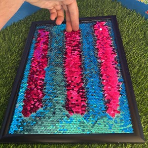 May include: A framed sensory toy with a black border. The interior features a background of blue and teal sequins, with three vertical columns of pink sequins. The sequins change colour when touched.