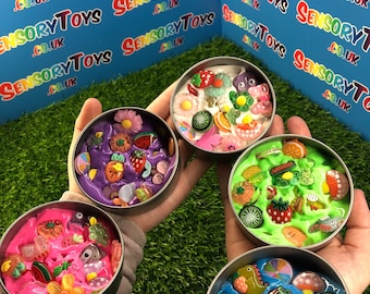 May include: Four small round containers filled with colorful slime and various small plastic charms. The containers are on a green grassy surface.