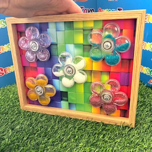 May include: A wooden-framed sensory toy with five clear, flower-shaped spinners filled with glitter and beads. The background is a mosaic of colourful blocks. The toy is designed for sensory exploration and play.