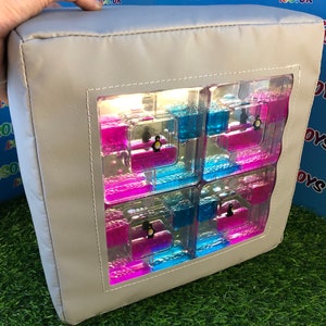 Könnte beinhalten: Ein graues Sinneskissen mit einem durchsichtigen Fenster, das ein flüssiges Sinnesspielzeug enthüllt. Das Spielzeug zeigt eine blaue und rosa Flüssigkeit mit kleinen Pinguinen, die darin schwimmen.
