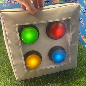 Puede incluir: Un cubo sensorial gris con cuatro botones iluminados de colores: verde, rojo, amarillo y azul. Los botones están dentro de una ventana transparente. El cubo es sostenido por una mano.