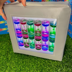 Könnte beinhalten: Ein grauer Sensorikwürfel mit einem transparenten Fenster, das farbenfrohe Flüssigkeitstimer zeigt. Die Timer enthalten blaue, rosa und grüne Flüssigkeiten. Der Würfel wird von einer Hand gehalten und liegt auf einer grünen Oberfläche.