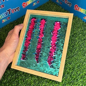 Op de afbeelding: Een sensorisch speeltje met een houten frame. De binnenkant heeft een turquoise achtergrond bedekt met pailletten. Drie verticale kolommen roze pailletten zijn zichtbaar. Het speelgoed is ontworpen voor tactiele verkenning en sensorisch spel.