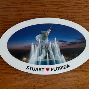 May include: Oval-shaped souvenir sticker featuring a vibrant image of a sailfish leaping above a fountain. The background shows a twilight sky. The text "STUART ❤️ FLORIDA" is printed at the bottom.