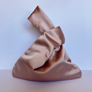 May include: A blush-colored satin handbag with a knotted handle. The bag has a soft, flowing shape and a subtle sheen. The background is a gradient of white and light pink.