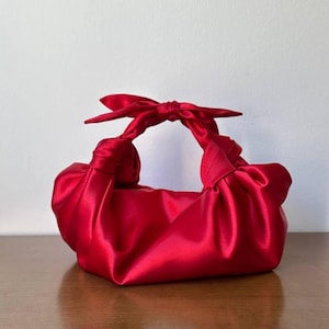 Pode incluir: Uma bolsa de mão em cetim vermelho vibrante com uma estrutura de tecido macio e franzido. A bolsa é projetada com duas alças com nós que se encontram no topo, criando uma aparência de laço. O material possui um brilho suave e reflexivo.