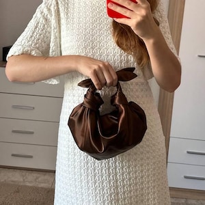 May include: A brown satin handbag with knotted handles. The bag is held in front of a person wearing a white textured dress. The person is holding a red phone in their other hand. The background includes a white dresser and a light-colored wall.