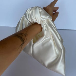 May include: Ivory satin clutch bag with a knotted handle. The bag is made of a soft, flowing fabric and has a simple, elegant design. The clutch is held by a hand.