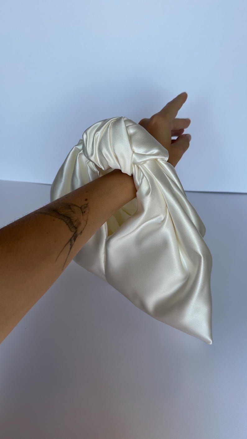 May include: Ivory satin clutch bag with a knotted handle. The bag is made of a soft, flowing fabric and has a simple, elegant design. The clutch is held by a hand.