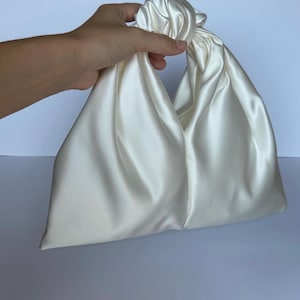 May include: Ivory satin handbag with a knotted handle. The bag has a triangular shape and a soft, flowing texture. The handle is formed by twisting the fabric, creating a unique and elegant design. The bag is held by a hand.