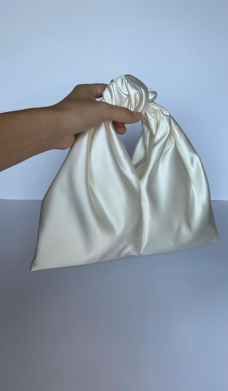 May include: Ivory satin handbag with a knotted handle. The bag has a triangular shape and a soft, flowing texture. The handle is formed by twisting the fabric, creating a unique and elegant design. The bag is held by a hand.