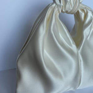 May include: Ivory satin handbag with a knotted handle. The bag has a soft, draped design and a smooth, lustrous finish. The handle is formed by a large knot, creating a unique and elegant look. The bag is a neutral color, suitable for various occasions.