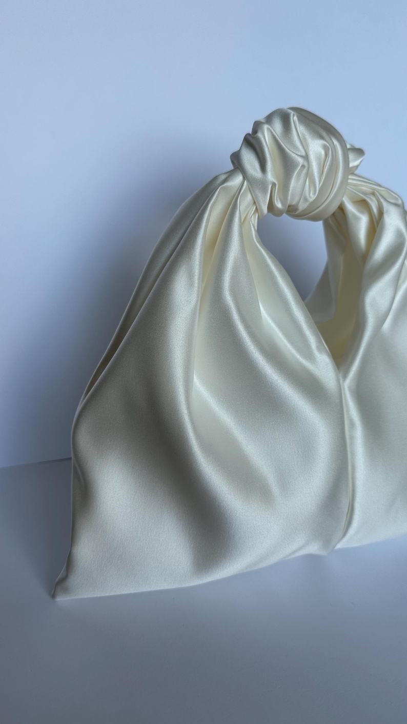 May include: Ivory satin handbag with a knotted handle. The bag has a soft, draped design and a smooth, lustrous finish. The handle is formed by a large knot, creating a unique and elegant look. The bag is a neutral color, suitable for various occasions.