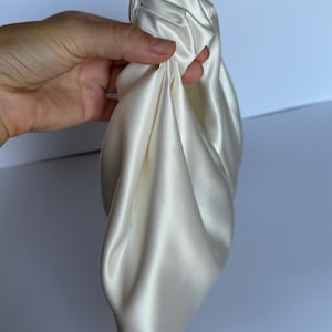 May include: Ivory-colored satin clutch bag, held in a hand. The bag is gathered and knotted at the top, with soft folds of fabric. The satin has a subtle sheen, reflecting light. The background is a plain white.