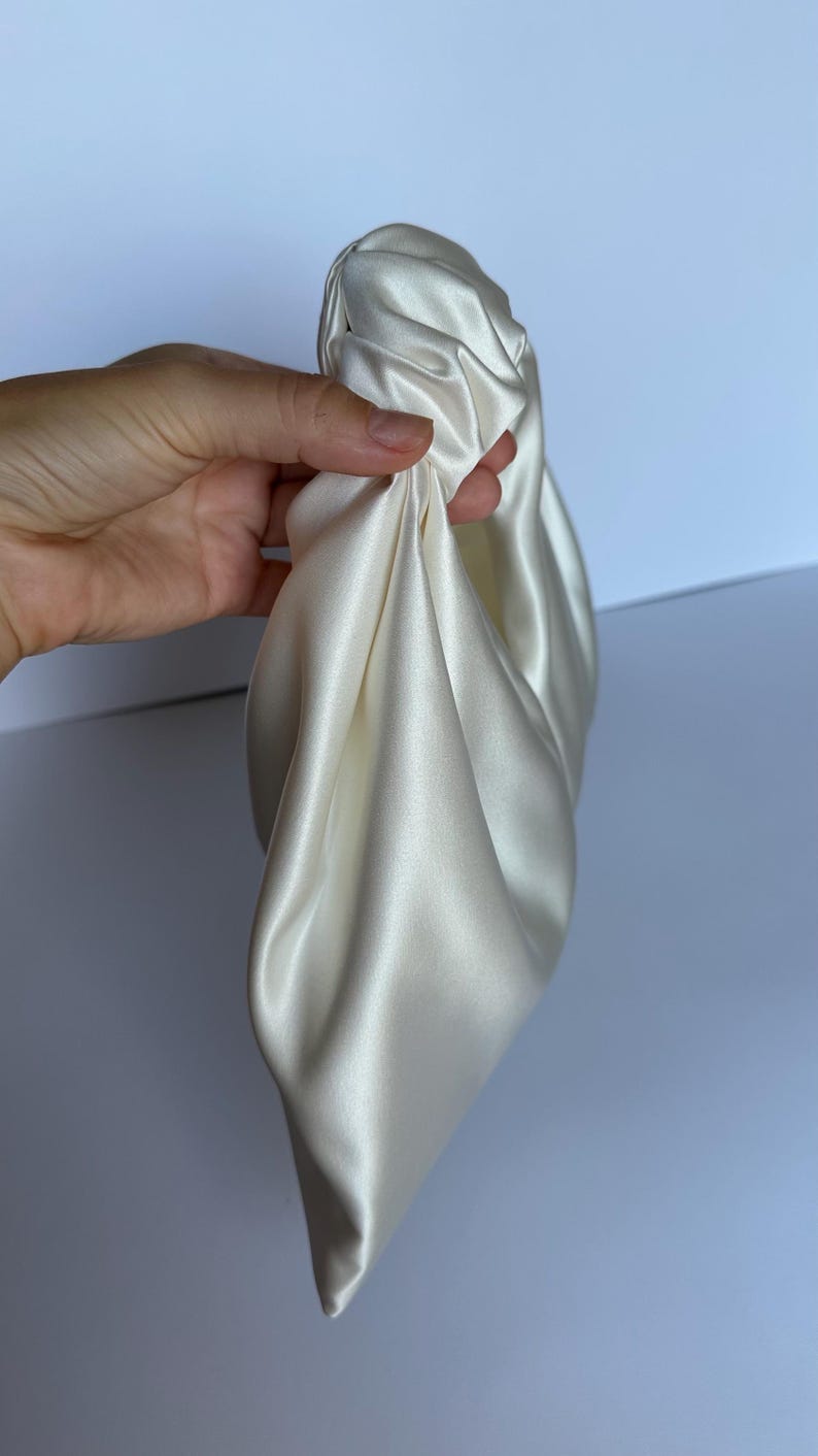 May include: Ivory-colored satin clutch bag, held in a hand. The bag is gathered and knotted at the top, with soft folds of fabric. The satin has a subtle sheen, reflecting light. The background is a plain white.